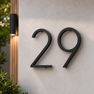 May include: A set of two large, matte black house numbers displaying the digits 2 and 9. The numbers feature a clean, sans-serif typeface with a three-dimensional profile, mounted directly onto a light-colored, textured exterior wall surface.