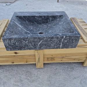 May include: A square bathroom sink carved from dark gray marble with prominent white veining. The basin features a deep, sloped interior with a central drain hole. The sink is positioned on a light-colored wooden shipping crate with slats.