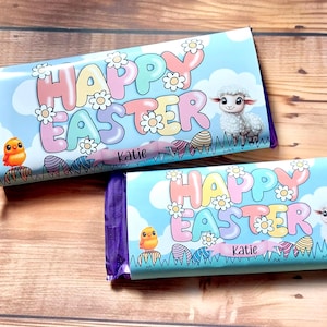 Pode incluir: Duas barras de chocolate embrulhadas em roxo com um fundo azul e letras em cores pastel que soletram "Feliz Páscoa". As barras apresentam um pintinho e um cordeiro, e o nome "Katie" está impresso em cada barra.