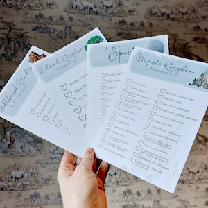 May include: A hand holds four white paper checklists with ride names for Disney theme parks. The checklists are titled "Hollywood Studio," "Animal Kingdom," "Epcot," and "Magic Kingdom." Each list has a light blue watercolor wash background.