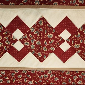 May include: Close-up of a quilted fabric with a repeating pattern. The design features alternating sections of floral print and solid cream-colored shapes. The floral print is a deep red with blue and tan flowers.