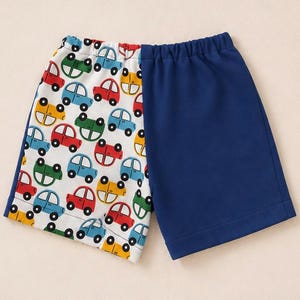 Op de afbeelding: Shorts met een gespleten ontwerp, één kant in effen marineblauw en de andere kant in witte stof met een patroon van kleine, kleurrijke cartoonauto's in rood, blauw, groen en geel. Het kledingstuk heeft een elastische tailleband en een zoom.