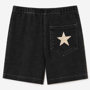 Op de afbeelding: Zwarte denim shorts met een elastische tailleband en een enkele achterzak. De zak is versierd met een grote, gecentreerde, gebroken witte ster met een textuur en gerafelde randen. De stof heeft een gewassen, vervaagde zwarte afwerking.