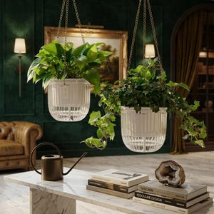 May include: Two clear, ribbed glass hanging planters suspended by gold chains, each containing a lush green plant. Below, a bronze watering can sits on a white marble table next to a stack of books and a geode, with a leather sofa in the background.