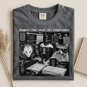 May include: A charcoal grey crew neck t-shirt featuring a graphic print of an opossum, a skunk, and a raccoon wearing headsets at a dispatch desk. The text reads Support Your Local 911 Dispatchers, Dispatch Saves Lives, and Fueled by Caffeine & Helping.