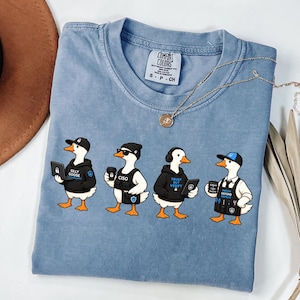 May include: A blue short-sleeved t-shirt featuring a graphic of four cartoon geese dressed as cybersecurity professionals. The geese wear hats, hoodies, and vests with text including Silly Goose, CISO, Trust But Verify, and Patch Defend Repeat. The shirt has a Comfort Colors brand label.