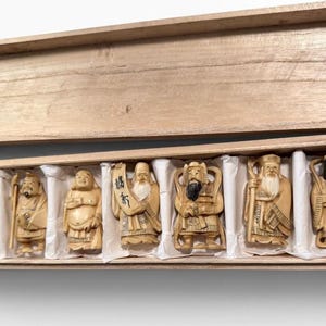 May include: A set of seven carved light-colored figurines depicting various figures, including deities and scholars, arranged in individual white paper-lined compartments within a rectangular, light-toned wooden box with a matching lid.
