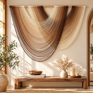 May include: A large wall hanging featuring multiple layers of draped cord in a gradient of cream, tan, and dark brown colors. The cords are suspended from a horizontal wooden rod, creating a series of overlapping U-shaped curves across the wall.
