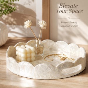 May include: A round, scalloped-edge tray with a pearlescent finish and integrated handles. On the tray sits a white bubble candle, a glass reed diffuser with two dried floral stems, and a delicate gold chain necklace. Text reads Elevate Your Space.