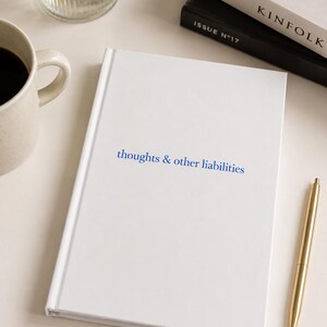 以下が含まれることがあります： 表紙に青色で thoughts & other liabilities と印刷された白いハードカバーのジャーナル。ジャーナルの横には、金色の金属製ペン、暗い液体が入ったセラミック製のマグカップ、白黒の表紙の積み重ねられた2冊の本が置かれています。