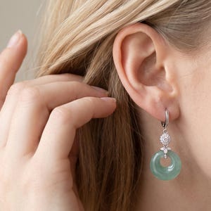 Puede incluir: Un pendiente colgante de tono plateado que presenta un colgante circular de piedra translúcida de color verde claro similar al jade, suspendido de una pequeña pieza conectora redonda con piedras transparentes y un cierre de aro.