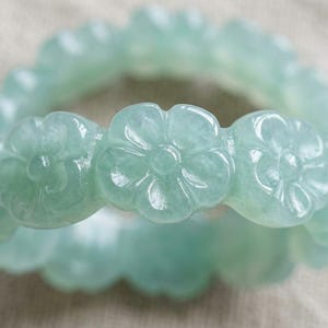May include: A pale green jade ring featuring a continuous band of carved floral motifs. The surface of the ring is smooth and polished, showcasing a translucent quality with subtle variations in the light green color throughout the flower shapes.