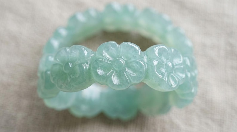 May include: A pale green jade ring featuring a continuous band of carved floral motifs. The surface of the ring is smooth and polished, showcasing a translucent quality with subtle variations in the light green color throughout the flower shapes.
