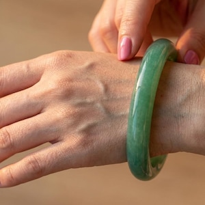 Może przedstawiać: Gładka, polerowana, cylindryczna bransoleta typu bangle wykonana z litego kawałka półprzezroczystego zielonego kamienia. Bransoleta ma zaokrąglony profil z błyszczącym wykończeniem, ukazującym naturalne różnice w jasnych i ciemnych odcieniach zieleni.