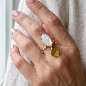 Puede incluir: Un anillo dorado con una incrustación de nácar blanco en forma de marquesa colocada verticalmente sobre un pequeño disco dorado circular y liso. El aro del anillo es fino y pulido, creando un diseño equilibrado con formas geométricas.