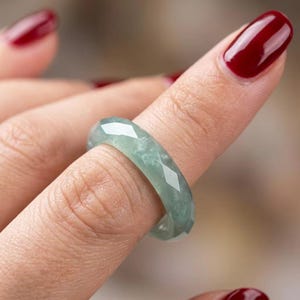 Puede incluir: Un anillo facetado elaborado con una piedra verde pálido translúcida con sutiles inclusiones blancas nubladas. La banda presenta un corte geométrico de múltiples lados que crea una serie de superficies planas y reflectantes alrededor.