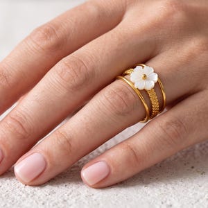 May include: A gold-toned multi-band ring featuring a white flower-shaped accent with a small gold centre bead. The ring consists of three distinct bands, one of which has a textured, beaded surface, while the other two are smooth and plain.
