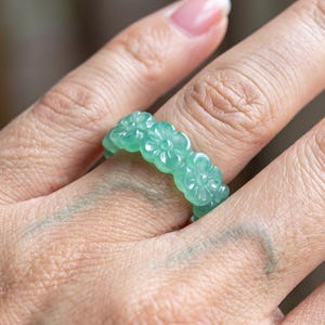 May include: A ring crafted from a single piece of translucent light green stone, featuring a continuous band of carved floral motifs. The design consists of multiple connected flower shapes with detailed petals encircling the entire circumference.