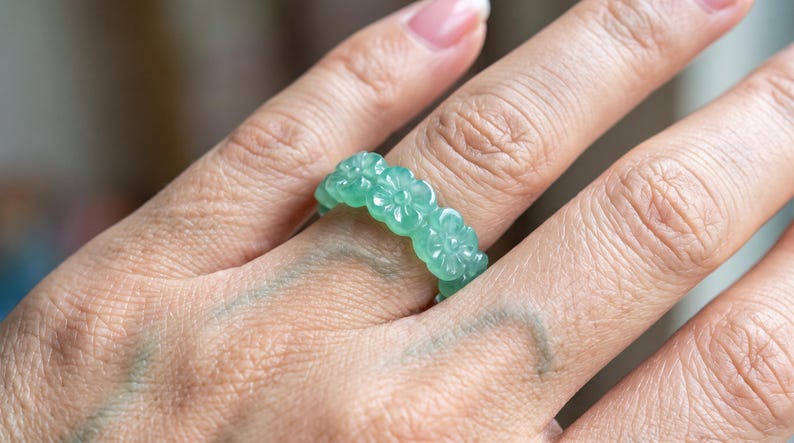 May include: A ring crafted from a single piece of translucent light green stone, featuring a continuous band of carved floral motifs. The design consists of multiple connected flower shapes with detailed petals encircling the entire circumference.
