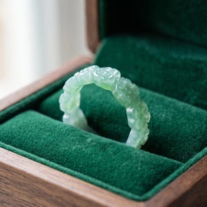 May include: A pale green, translucent ring carved with a repeating floral pattern along the entire band. The ring is positioned upright within a dark brown wooden jewelry box featuring a plush, deep green velvet interior lining.