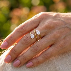 Puede incluir: Un anillo de oro tipo bypass con dos piedras claras de corte ovalado montadas sobre una banda fina y pulida. El anillo presenta un espacio abierto entre las dos piedras, situadas en el dedo índice y el corazón, con un diseño sencillo.