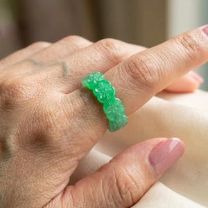 May include: A bright green ring carved from a translucent stone material, featuring a continuous band of repeating floral motifs. The ring has a smooth, polished finish and a vibrant, semi-transparent appearance that catches the light.