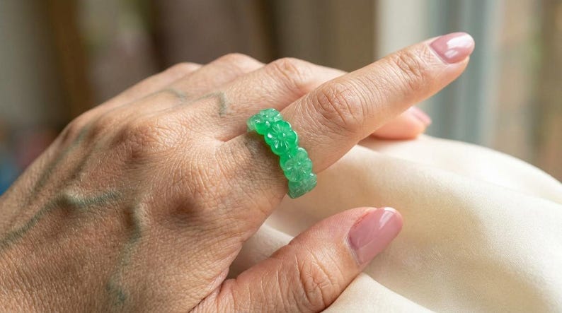 May include: A bright green ring carved from a translucent stone material, featuring a continuous band of repeating floral motifs. The ring has a smooth, polished finish and a vibrant, semi-transparent appearance that catches the light.