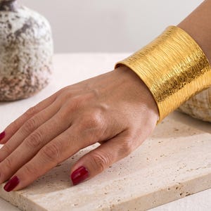 Puede incluir: Un brazalete ancho de oro con un acabado de metal cepillado y texturizado. La pulsera tiene una superficie pulida y reflectante con finas líneas horizontales grabadas en toda la banda, creando un patrón consistente y repetitivo.