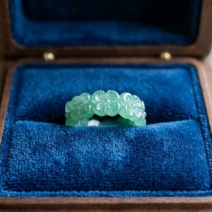 May include: A light green translucent ring carved with a repeating floral pattern, featuring five-petaled blossoms encircling the entire band. The ring is displayed inside a dark wooden box lined with deep blue velvet fabric.