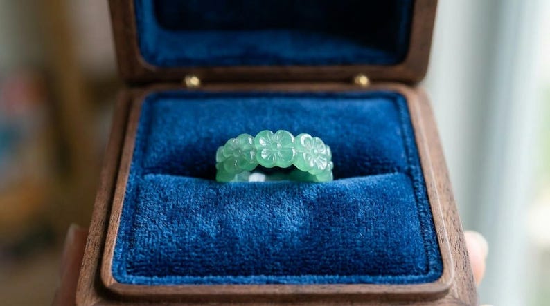 May include: A light green translucent ring carved with a repeating floral pattern, featuring five-petaled blossoms encircling the entire band. The ring is displayed inside a dark wooden box lined with deep blue velvet fabric.