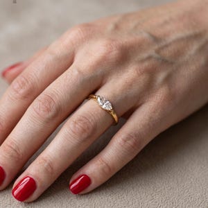 Puede incluir: Un anillo de oro con una sola piedra preciosa blanca de talla marquesa engastada horizontalmente en una banda de oro lisa y pulida. El anillo se lleva en un dedo, destacando la forma alargada de la piedra y el brillo del metal dorado.