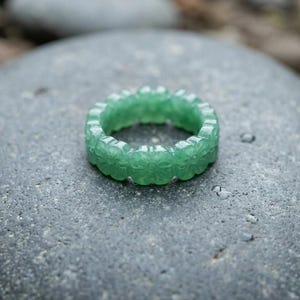 May include: A green ring carved from a single piece of stone, featuring a repeating floral or clover-like pattern around the entire band. The ring has a smooth, polished finish and a translucent quality, showcasing a vibrant, consistent emerald green hue.