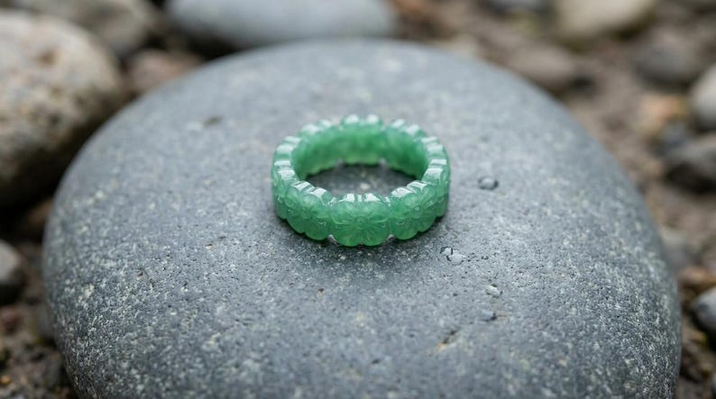 May include: A green ring carved from a single piece of stone, featuring a repeating floral or clover-like pattern around the entire band. The ring has a smooth, polished finish and a translucent quality, showcasing a vibrant, consistent emerald green hue.