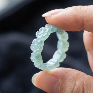 May include: A pale green, translucent ring carved from a single piece of stone. The band features a repeating pattern of small, five-petaled flowers encircling the entire circumference, showcasing a smooth, polished finish and subtle color variations.
