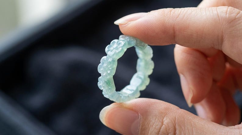 May include: A pale green, translucent ring carved from a single piece of stone. The band features a repeating pattern of small, five-petaled flowers encircling the entire circumference, showcasing a smooth, polished finish and subtle color variations.