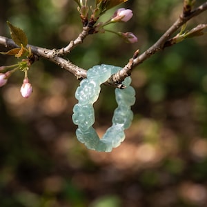 May include: A pale green, translucent ring carved from a single piece of stone. The band features a continuous pattern of small, five-petaled flowers linked together in a circular shape, creating a smooth, polished surface throughout the design.