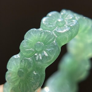 May include: A light green translucent ring carved from a single piece of stone. The band features a continuous pattern of relief-carved five-petaled flowers with central circular details, creating a textured surface around the entire circumference.
