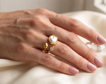 May include: An open-style gold ring featuring a large, irregular white pearl on one end and a small, round, clear gemstone on the other. The band has a textured, organic finish, and the design is worn on a finger with light pink nail polish.