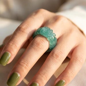 Puede incluir: Un anillo de color verde azulado translúcido con un patrón floral de rosas tallado que envuelve toda la banda. El anillo tiene un acabado liso y pulido con un perfil redondeado, mostrando detalles intrincados de pétalos tallados en el material.