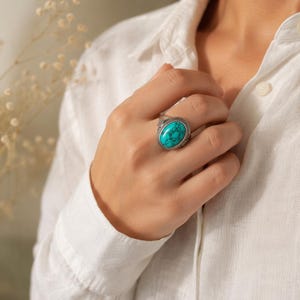 Puede incluir: Un anillo de plata con una gran piedra turquesa de forma ovalada con vetas oscuras. La banda de metal presenta detalles texturizados intrincados alrededor del engaste, proporcionando un marco decorativo para la gema azul verdosa.