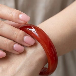 Puede incluir: Una pulsera tipo brazalete de color rojo intenso, lisa y pulida, con un perfil redondeado y acabado brillante. La banda circular sólida presenta un tono carmesí vibrante y constante en toda su estructura, reflejando la luz en su superficie.
