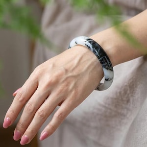 以下が含まれることがあります： 黒と白の渦巻き模様が特徴的な、滑らかで丸みを帯びたバングルブレスレット。円形のアクセサリーは光を反射する磨き上げられた光沢のある仕上げで、バンド全体に流れるようなデザインが施されています。