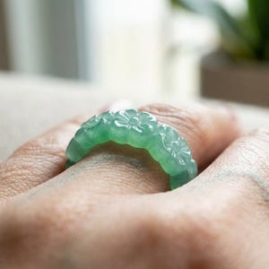 May include: A translucent light green ring carved from a single piece of stone. The band features a continuous pattern of small, raised floral motifs encircling the entire circumference, creating a textured and detailed surface design.