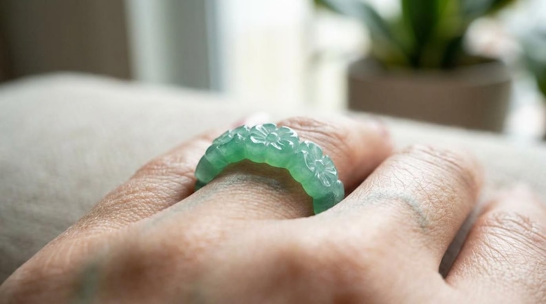 May include: A translucent light green ring carved from a single piece of stone. The band features a continuous pattern of small, raised floral motifs encircling the entire circumference, creating a textured and detailed surface design.