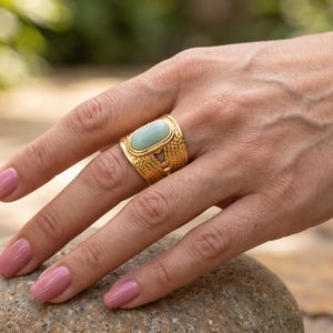 Puede incluir: Un anillo ancho de metal dorado con una piedra ovalada de color verde claro en el centro. La banda presenta un patrón texturizado de líneas entrecruzadas en toda su superficie, aportando un acabado detallado a la joya.