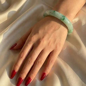 Puede incluir: Una pulsera tipo brazalete de jade pulido y liso, con una base de color verde pálido translúcido y distintas inclusiones moteadas de color verde más oscuro. El brazalete circular tiene un perfil redondeado y una superficie brillante.