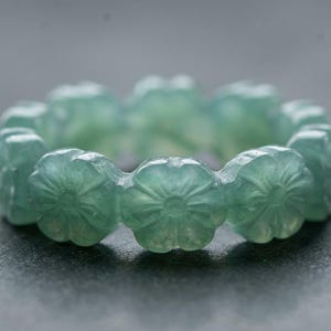 May include: A light green ring carved from a single piece of stone, featuring a continuous band of repeating flower motifs. The translucent material displays subtle variations in color and texture throughout the circular floral design.