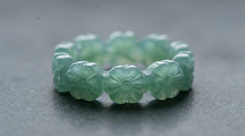 May include: A light green ring carved from a single piece of stone, featuring a continuous band of repeating flower motifs. The translucent material displays subtle variations in color and texture throughout the circular floral design.