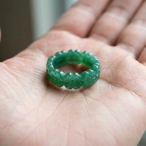 May include: A circular ring carved from a single piece of translucent green stone. The band features a repeating pattern of small, four-petaled flower shapes carved into the exterior surface, creating a scalloped edge around the entire circumference.