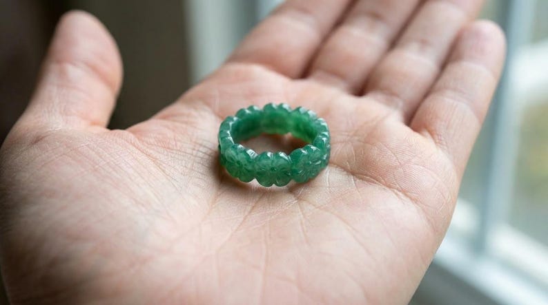 May include: A circular ring carved from a single piece of translucent green stone. The band features a repeating pattern of small, four-petaled flower shapes carved into the exterior surface, creating a scalloped edge around the entire circumference.